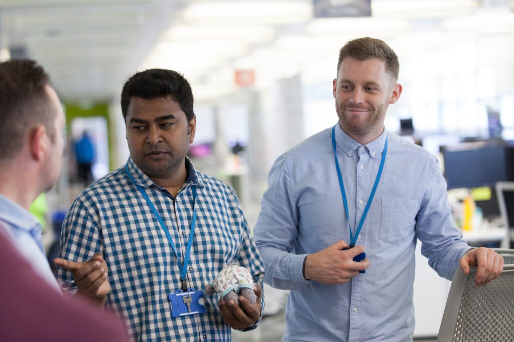 Three male staff standing in conversation in an open plan office