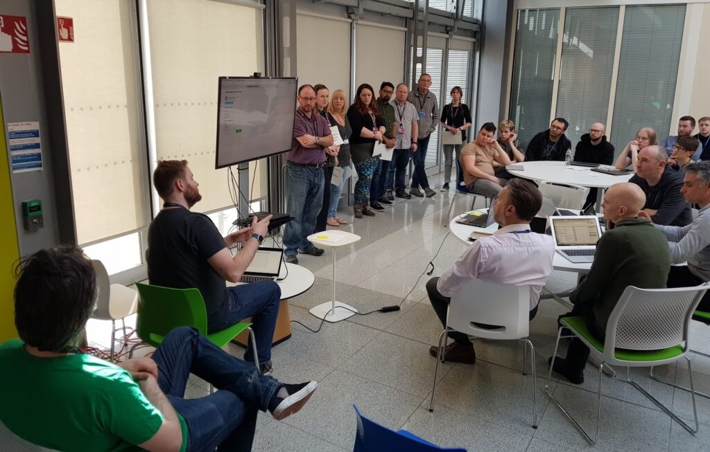 A team stand at the front of the seated audience and pitch their idea on a large screen