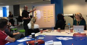 A male member of the network is standing at a whiteboard which is covered in yellow post it notes. He's talking to 5 delegates who are sitting around a table