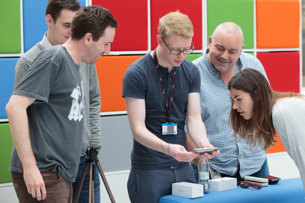 A group of five digital team members looking excitedly at a piece of tech kit in front of a brightly coloured background