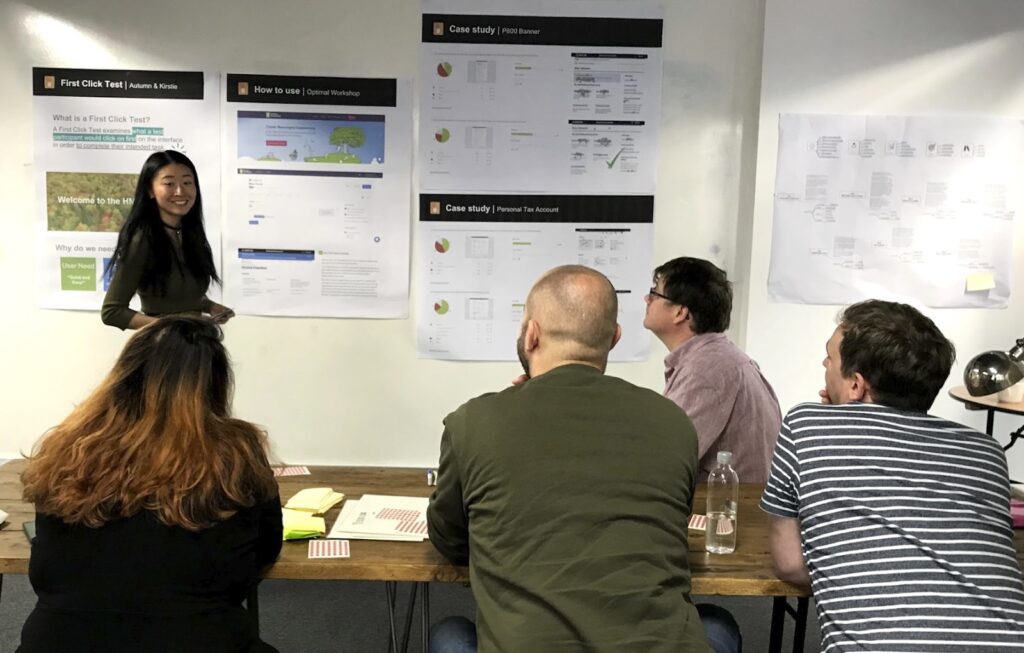User Research Autumn Wang is standing in front of a seated group with posters of the first click process behind her