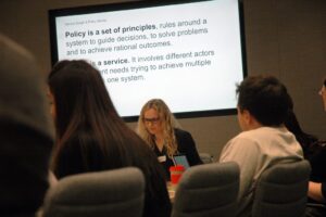 A large screen in the background with text about Policy Lab. People sitting around a conference table