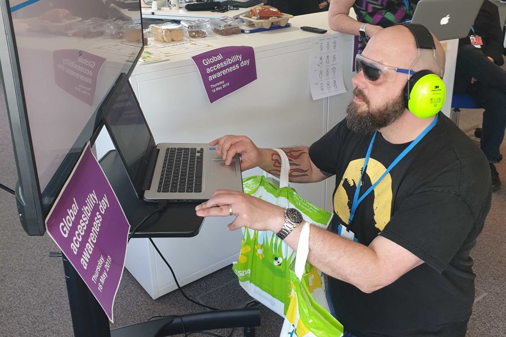 A man wearing blackout glasses and with his arms weighted by bags is trying to use an HMRC online service